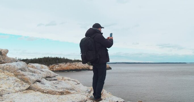 Young male Tourist standing on the top of a rocky mountain, photographing the ocean with a smartphone and admiring the panoramic sunset against the backdrop of the Atlantic Ocean.