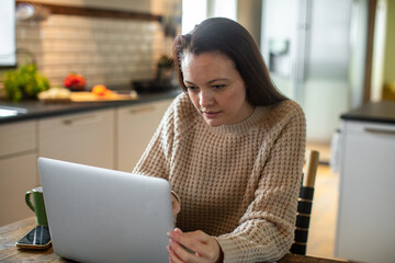 Fototapeta premium Young adult woman working on her laptop in the kitchen of her home