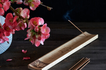 Burning japanese incense stick on wooden incense holder and vase with flowering chinese quince twigs on dark wooden table