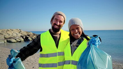 Happy multiracial volunteer couple showing the trash bag looking at camera after collect plastic waste from the sand of the beach. Eco activist people picking up and recycling rubbish from the nature