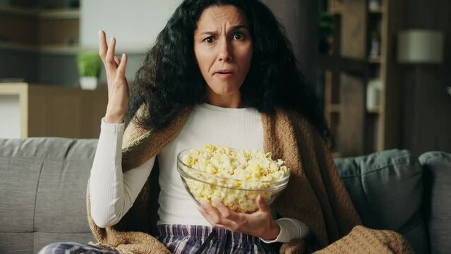 Camera view of angry hispanic woman looks at tv screen with negative emotion and throwing popcorn at the screen during leisure time at home Emotional female dislike boring film movie show indoors