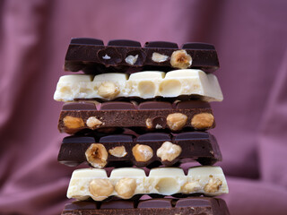 Stack of half chocolate bars with hazelnuts - milk chocolate, dark chocolate and white chocolate on a burgundy background.