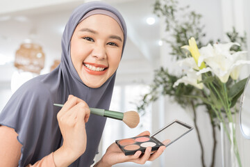 Asian young Muslim woman applying makeup with brush while looking in mirror. Asian Muslim female wear hijab headscarf applying cosmetic mascara on cheeks. Muslim woman makeup with blush on cheekbones