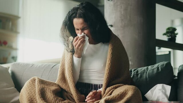 Sick hispanic young woman sitting on sofa under the warm blanket coughing sneezing and having severe headache temperature at home Unhealthy female getting flu virus symptom Cold and fever concept