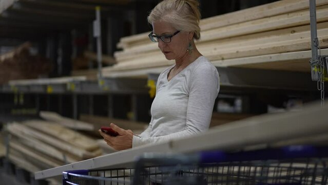 Attractive mature elderly blonde woman using smart phone in lumber section of hardware store. Concept of older person shopping for home improvement experience
