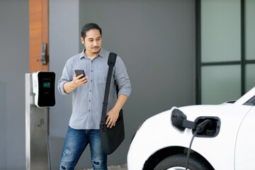 Progressive asian man and electric car with home charging station. Concept of the use of electric...