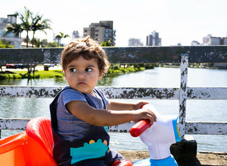Baby sitting on a blue and red scooter on a bridge over a lake in a quaint urban location with animals, trees and buildings. Curiosity and new learning outdoors and neurological development.