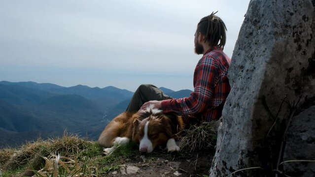 Travel concept. Caucasian man with dreadlocks sitting on top of mountain and resting during hike with pet. The guy traveling with dog Australian Shepherd. Petting and hugging dog.