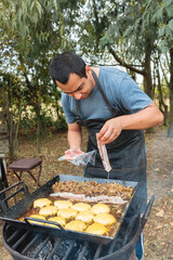Front view of a latin young man putting bacon and cheese on the burger on a barbecue grill