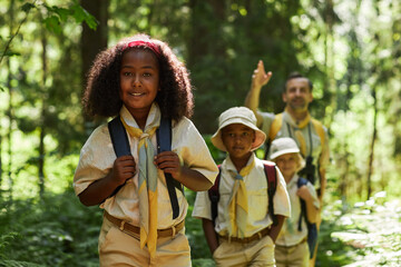 Waist up portrait of black girl with group of scouts hiking in forest and smiling at camera, copy space