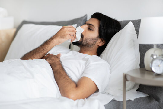 Sad young middle eastern guy in white t-shirt lies on bed, blows nose in napkin, sick and suffers from flu and cold
