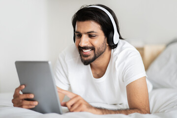 Glad young middle eastern man in white t-shirt and headphones typing on tablet, lies on bed, enjoys music