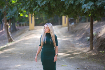 Beautiful blonde woman with straight hair dressed in a green dress walks through a field surrounded by trees and vegetation. The woman dresses elegantly and poses for the photo.