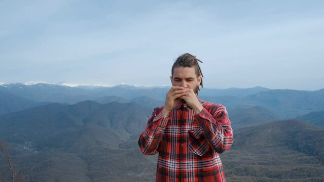 Concept of travel tourism and active healthy lifestyle. Caucasian man with dreadlocks stands on top of mountain and plays melody on harmonica, closing his eyes from pleasure of tranquility and peace.