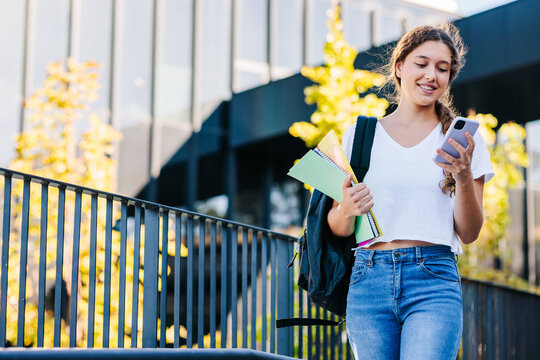 Smiling college student walking by her campus looking at her phone 