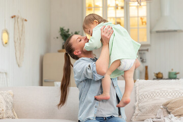 Happy family at home. Mother lifting in air little toddler child daughter. Mom and baby girl playing having fun together at home. Mother hugs baby with love care. Young mother kid rest in living room