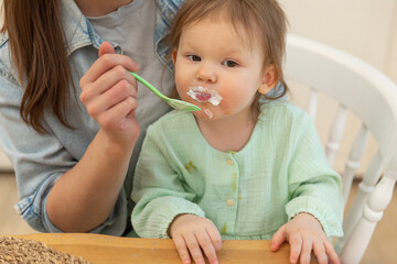 Happy family at home. Mother feeding her baby girl from spoon in kitchen. Little toddler child with messy funny face eats healthy food at home. Young woman mom giving food to kid daughter