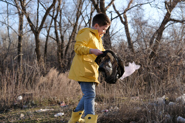 A cute little boy in yellow jacket and rubber boots is collecting garbage outside.
