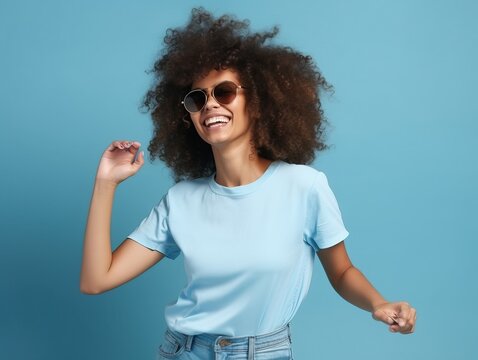 Gl&uuml;ckliche Frau mit Afrolocken tanzt in l&auml;ssigem T-Shirt und Sonnenbrille auf blauem Hintergrund, generative AI.