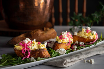 Creamy egg salad tops grilled slices of baguette on a bed of arugula. Garnished with pickled red onions and crumbled goat cheese.
