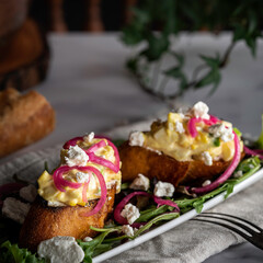 Creamy egg salad tops grilled slices of baguette on a bed of arugula. Garnished with pickled red onions and crumbled goat cheese.