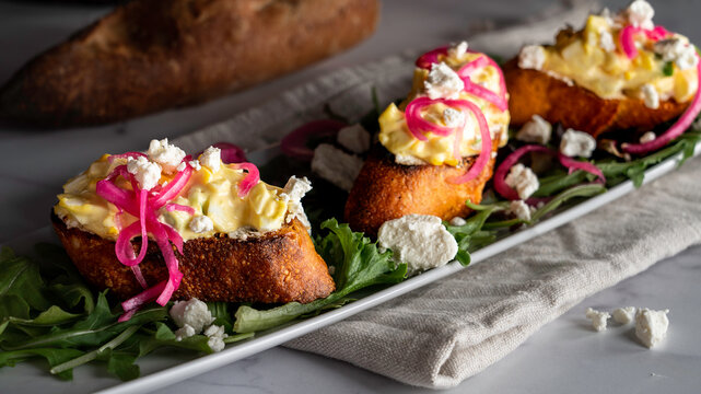 Creamy egg salad tops grilled slices of baguette on a bed of arugula. Garnished with pickled red onions and crumbled goat cheese.