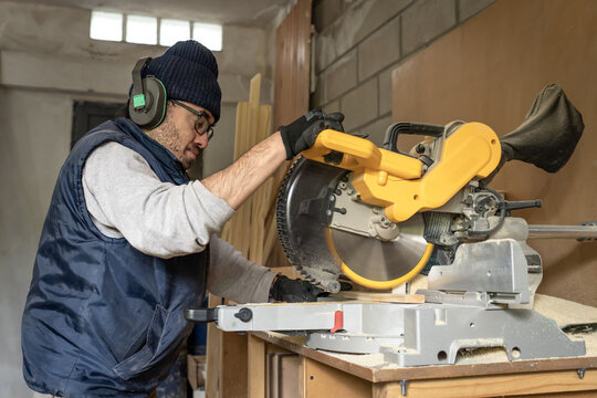 Carpenter Sawing Wood With An Electric Jigsaw In The Workshop With Ear Protection Material.