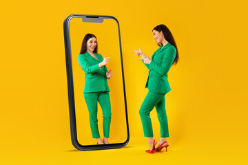Happy woman looking at giant cellphone display with her reflection, enjoying new outfit, standing on yellow background