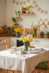 View of clean white simple modern scandinavian style kitchen, kitchen details, wooden table, bouquets of daffodils in vase on tables. Spring mood