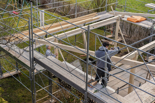Two young carpenters building a carport. One Carpenter drills a hole in a rafter with an electric drill, and stands on a railing. Second carpenter position a post.