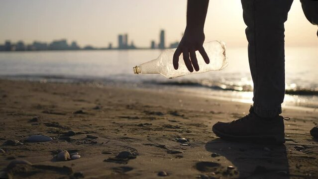 a male volunteer picks up plastic garbage at sunset against the background of a city on the seashore