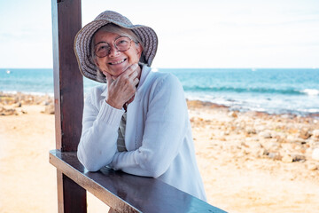 Smiling senior woman in casual clothing with hat and eyeglasses relaxed at the beach looking at camera enjoying vacations - horizon over sea