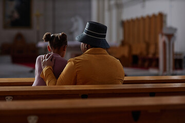 Rear view of grandmother visiting church with her granddaughter, they sitting on bench and praying...