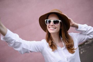 Young happy woman in summer clothes, sunglasses, straw hat posing on a pink background, taking a selfie portrait. Summer, technology concept