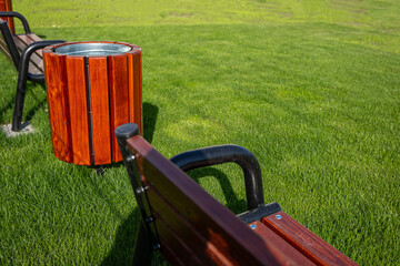 The trash can stands right next to a bench near the green lawn.