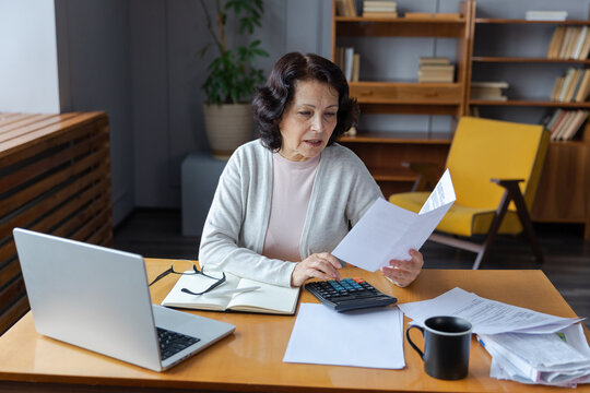 Middle Aged Senior Woman Sit With Laptop And Paper Document. Pensive Older Mature Lady Reading Paper Bill Pay Online At Home Managing Bank Finances Calculating Taxes Planning Loan Debt Pension Payment