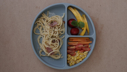 kids meal set, spaghetti carbonara with fruits and vegetables, carrot tomato and cord, on food grade plate for kid, children or toddler, top view