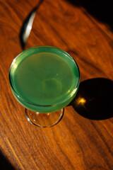 stirring green absinthe in a coupe glass with silver bar spoon on wooden table in the sun