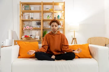 Wandcirkels plexiglas Yoga mindfulness meditation. Young healthy african girl practicing yoga at home. Woman sitting in lotus pose on couch meditating smiling relaxing indoor. Girl doing breathing practice. Yoga at home © Юлия Завалишина