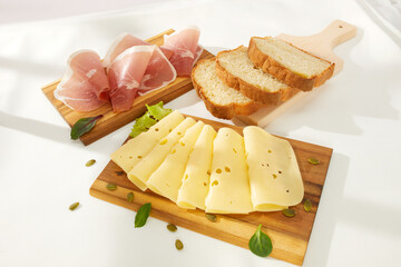 cheese slices with greens and prosciutto meat delicacy on a wooden board. bread is behind. snack on...