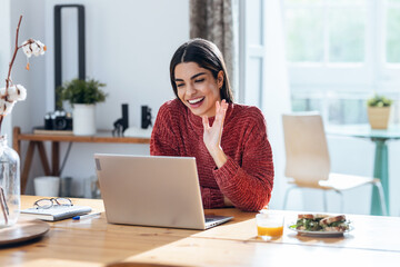 Beautiful young woman doing video call with laptop while having healthy sandwich for breakfast at home