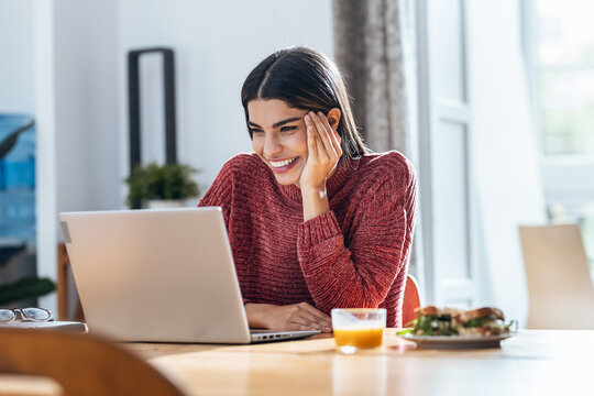 Beautiful young woman doing video call with laptop while having healthy sandwich for breakfast at home