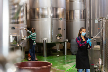 Portrait of female winery worker in face mask working with metal tanks for wine fermentation,...