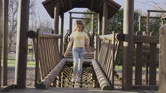 Little blonde girl walks concentrating on bridge of wooden playhouse on swings in park. Happy children people doing outdoor entertainment activities in spring on sunny day. Childhood and freedom.