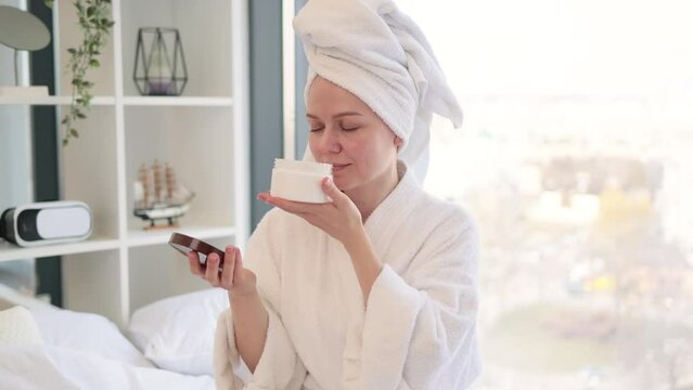 Close-up of lovely carefree lady in towel savouring pleasant fragrance of cosmetic product in hand while pampering herself with skincare procedures. Facial cream moisturizing skin after showering.