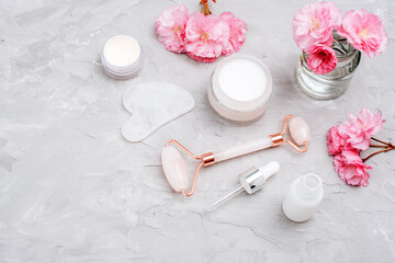 Cream jar, face roller, gua sha stone, serum bottle with pipette and pink cherry blossom on gray concrete background. Skin care, beauty treatment concept. Top view, flat lay