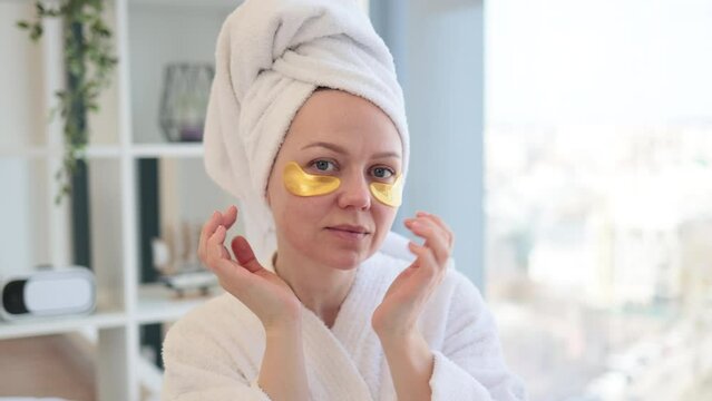 Close up view of lovely caucasian female in towel and dressing gown applying pair of eye patches after cleansing procedures in morning routine. Pretty woman taking care of delicate skin around eyes.