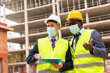 African-american and Eropean engineers in face masks standing on construction site and talking about new project. One man holding folder paper with documentation.