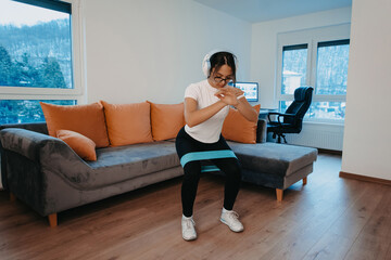 A young woman doing squats in a modern apartment, wearing headphones on her head, indicating she's listening to music or an audio workout guide. Her determined expression and active pose showcase her