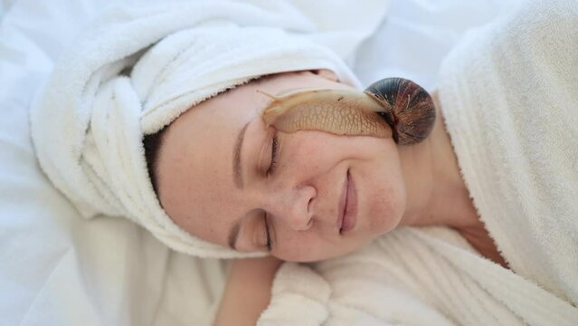 Close up view of young female lying with closed eyes on bed while cute mollusc slithering about on her face. Charming lady in soft towel enjoying snail facial procedure after light massage on bed.